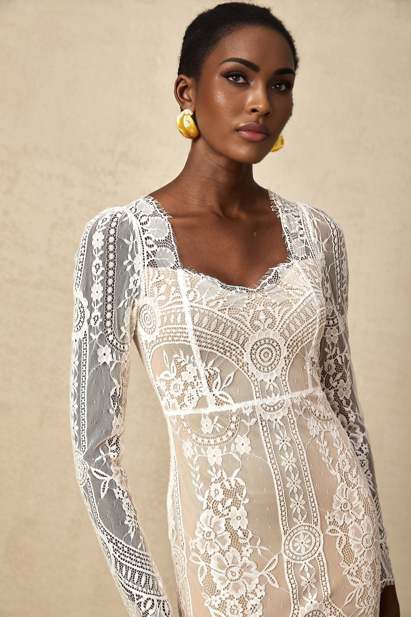 Woman wearing white lace mini dress with scalloped hem and short sleeves standing against plain background