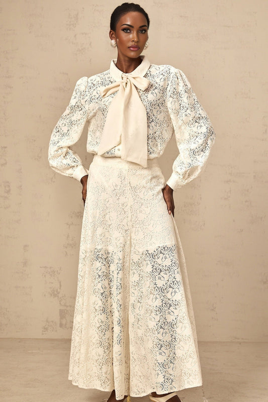 Woman wearing white lace shirt and matching trousers set against plain background