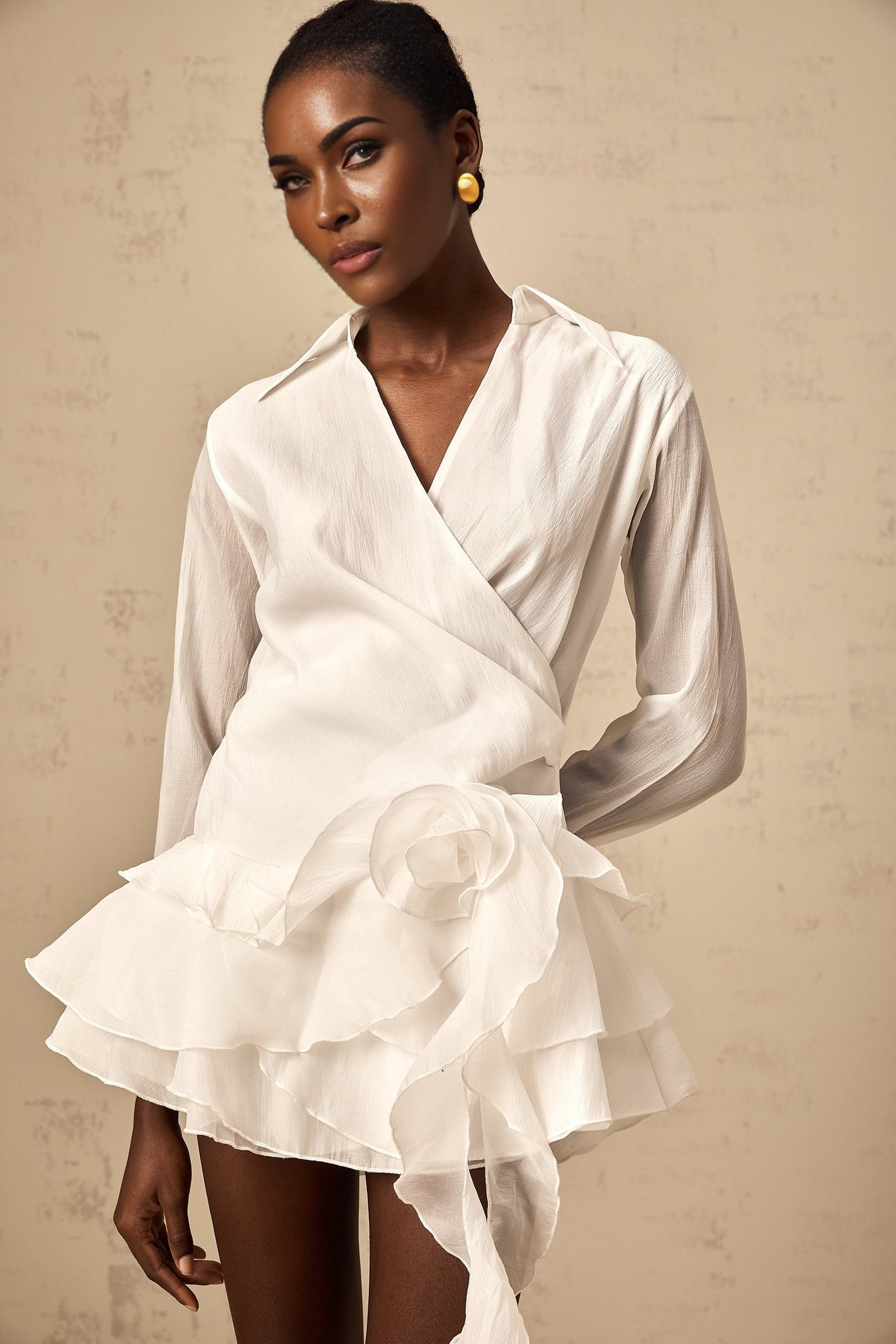 Woman wearing white semi sheer mini dress with floral pattern and ruffled details standing against plain background