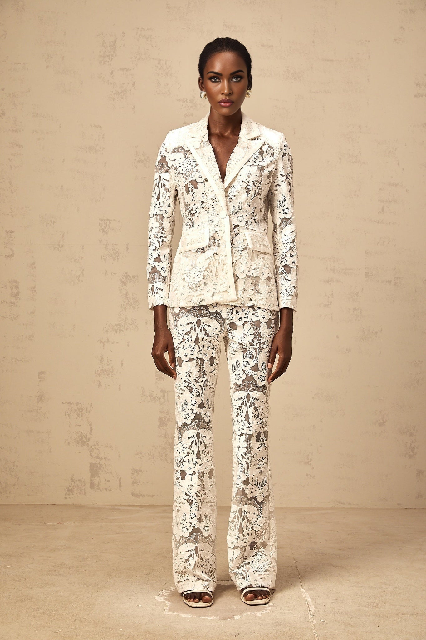 Woman wearing white tulle blazer and trousers set with sequined flower embroidery Yaelle matching outfit against plain background