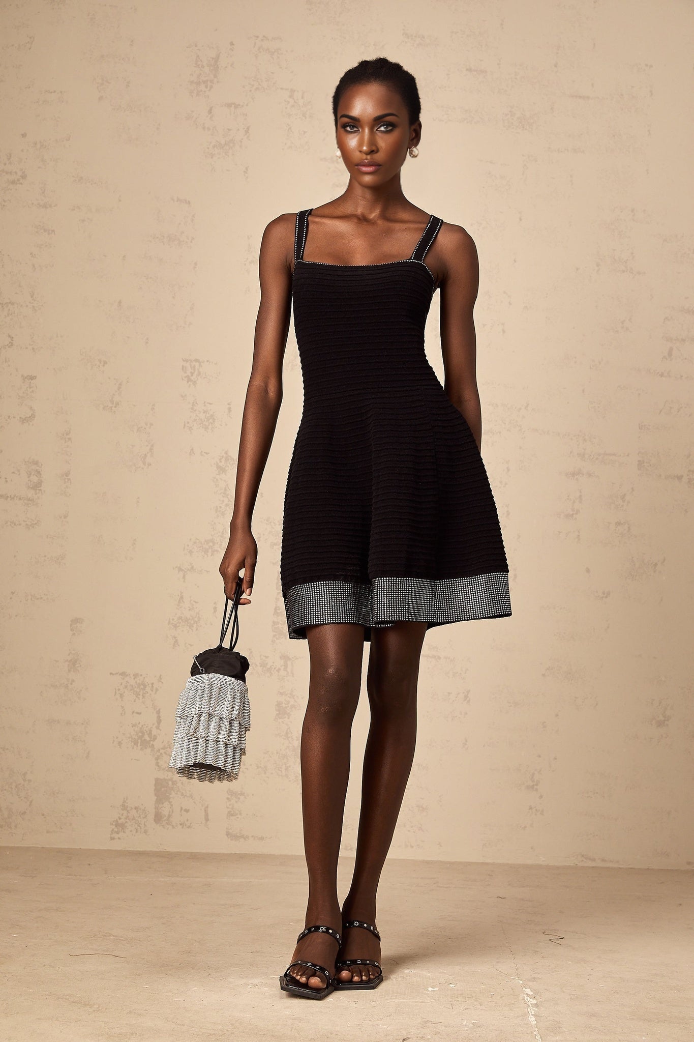 Young woman wearing a black knit mini dress with flared skirt and studded details standing in front of plain background
