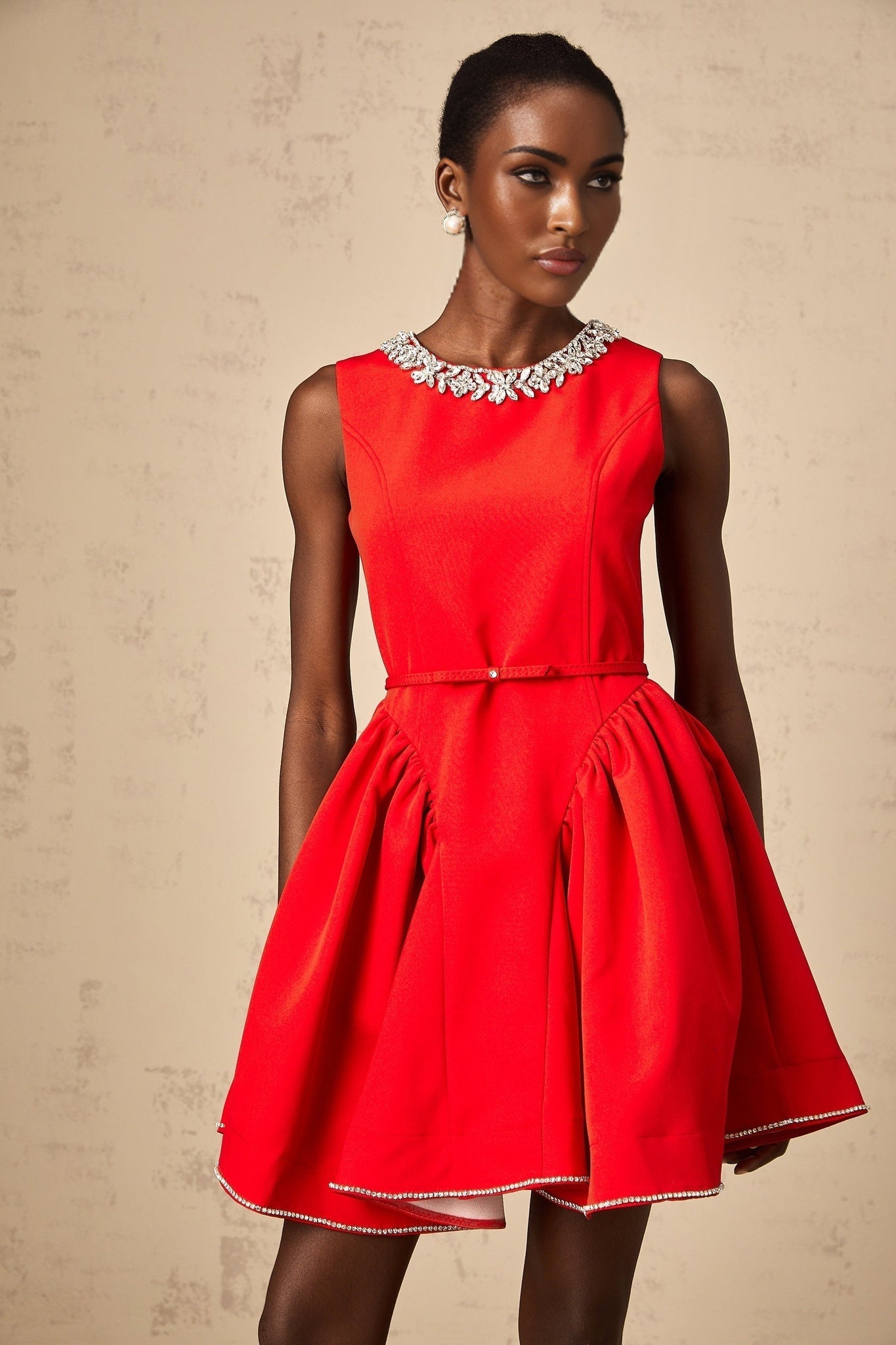 Young woman wearing a red mini dress with pleated skirt and embellished bodice standing in front of plain background
