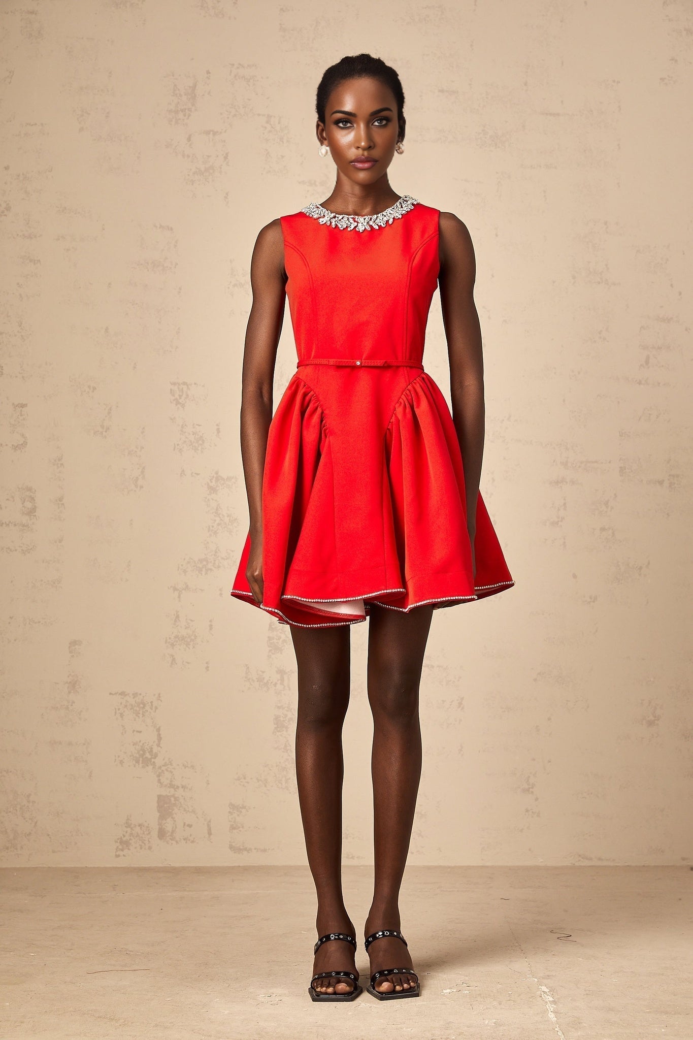 Young woman wearing a red mini dress with pleated skirt and embellished details standing in front of neutral background