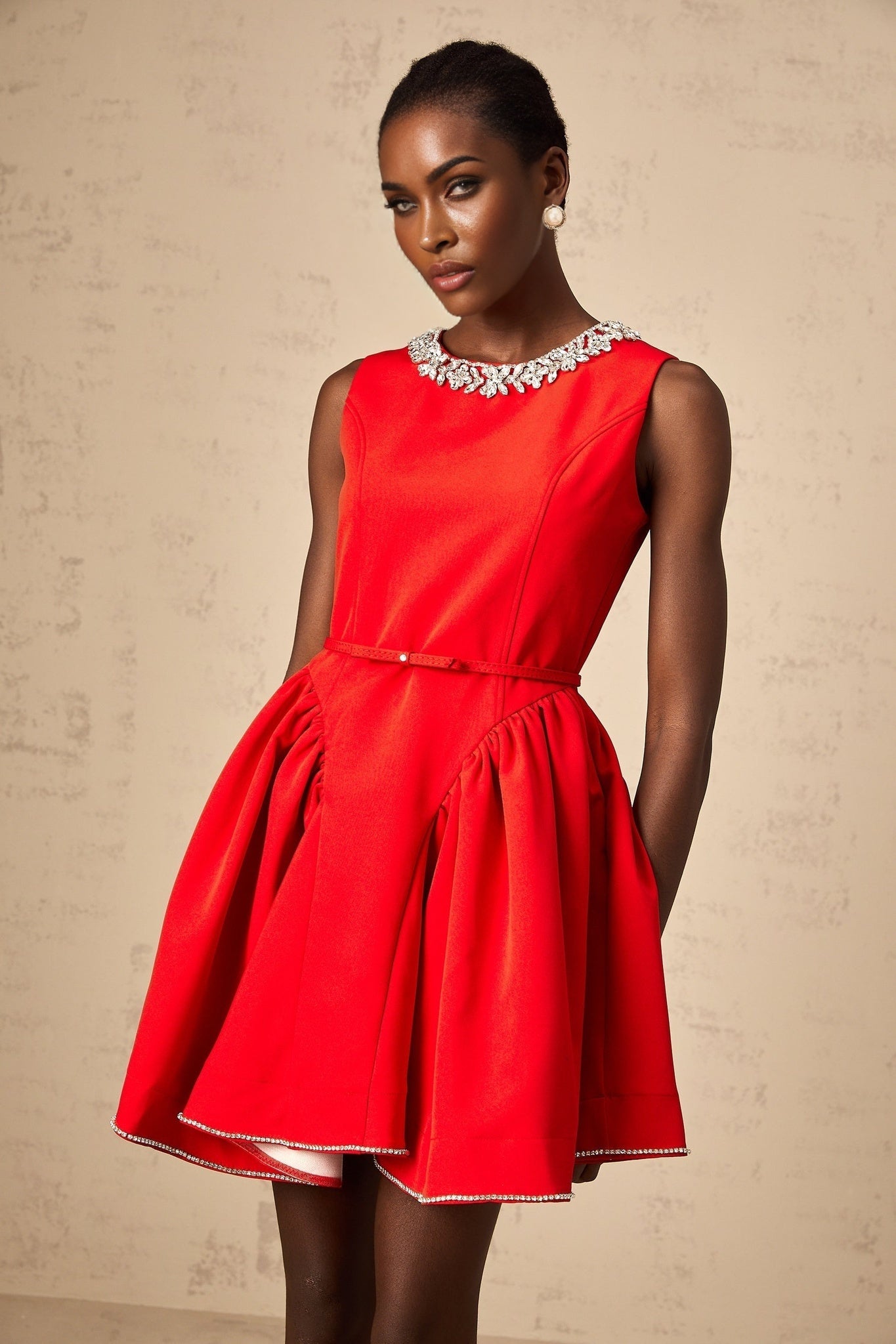 Young woman wearing a red pleated mini dress with embellishments standing against a plain background