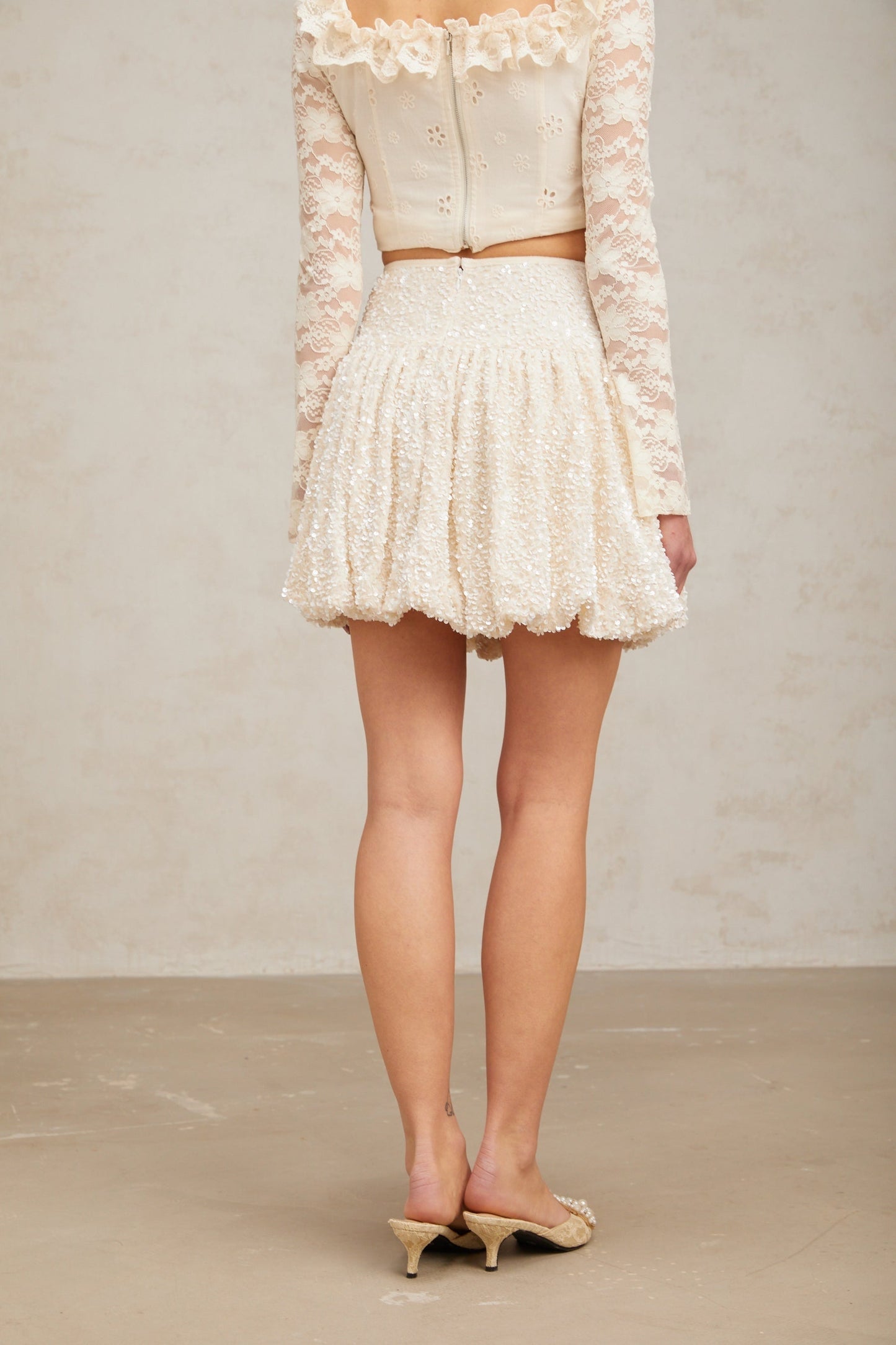 Young woman wearing a shimmering pleated mini skirt with sequin details standing against a plain background