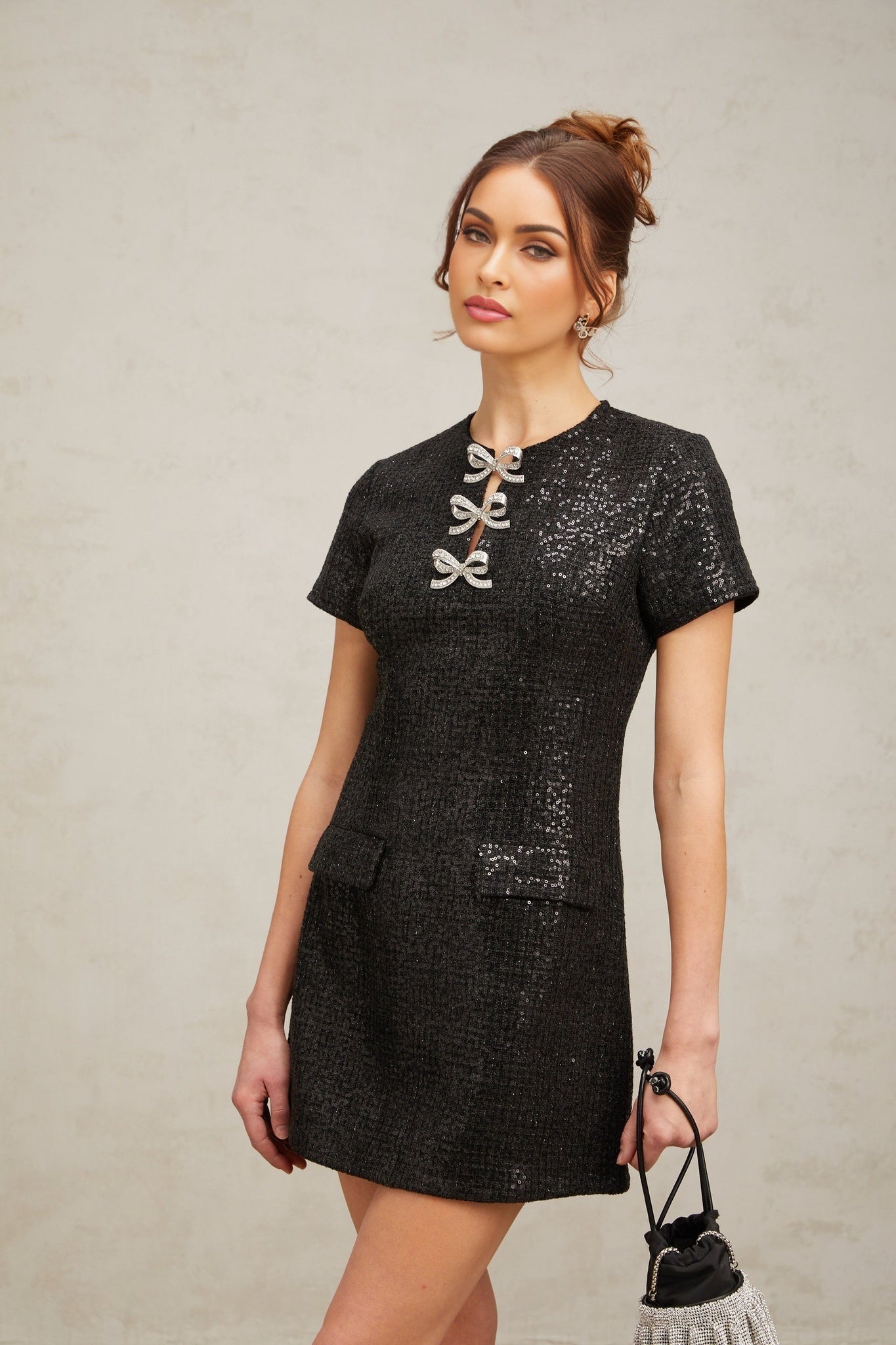Young woman wearing a short black mini dress with glitter details standing against a plain background