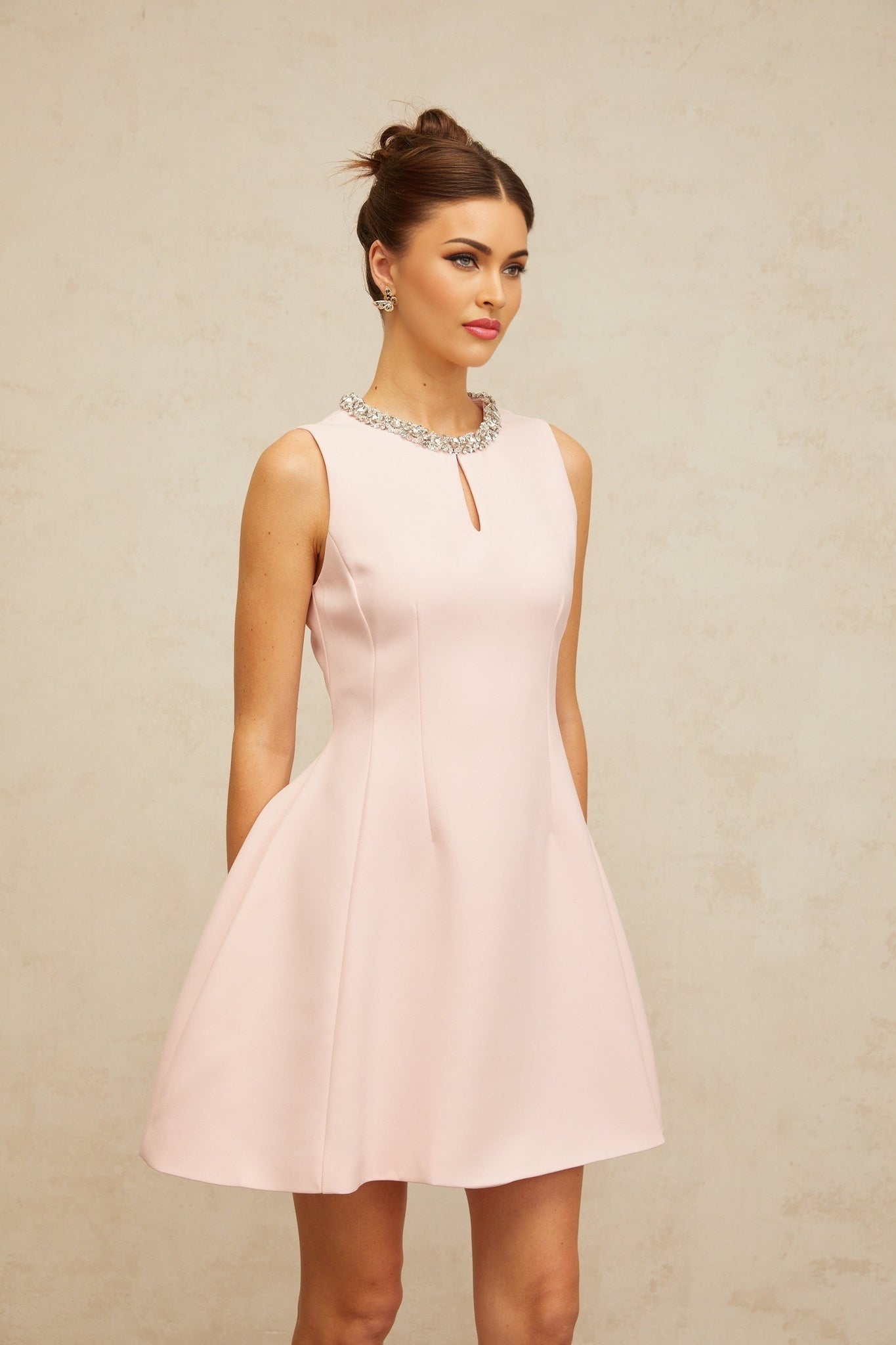 Young woman wearing a short pink mini dress with sequin embellishments and puff sleeves standing against a plain background