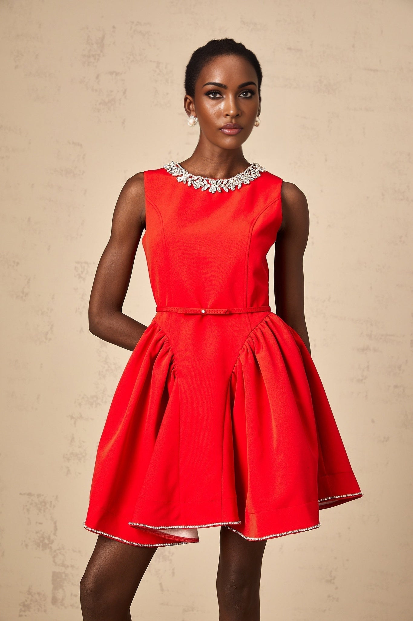 Young woman wearing a short red dress with pleated skirt and embellished details standing against a plain background