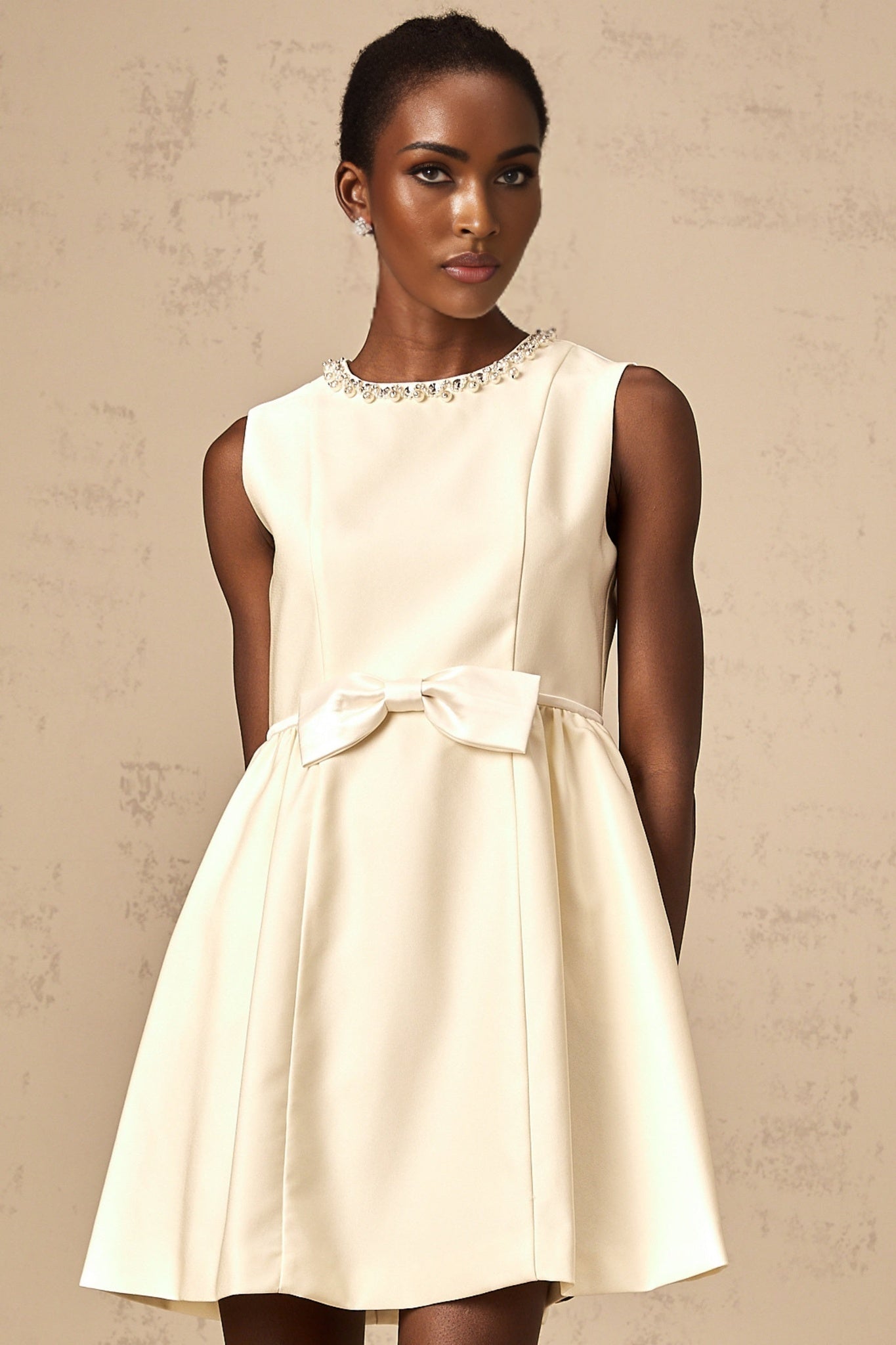 Young woman wearing a short white dress with a bowknot detail and embellishments standing against a plain background