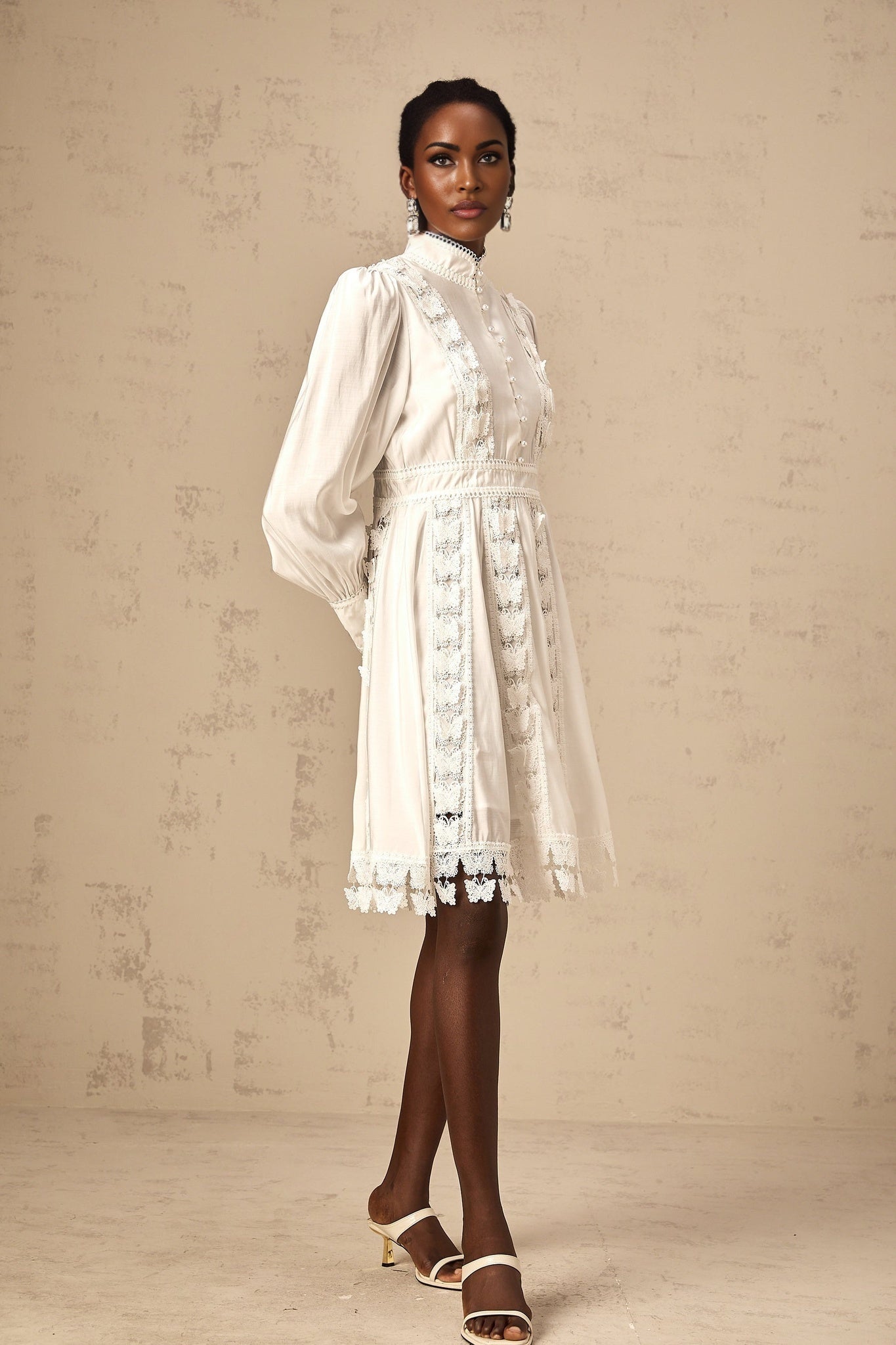 Young woman wearing a short white dress with butterfly appliques standing against a plain background