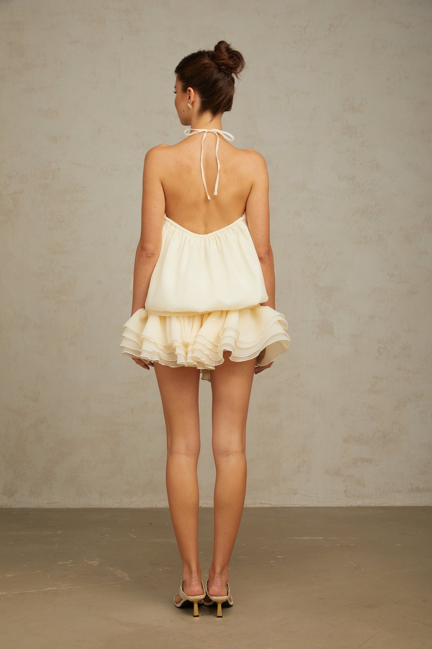 Young woman wearing a short white halterneck dress adorned with faux pearls standing against a plain background