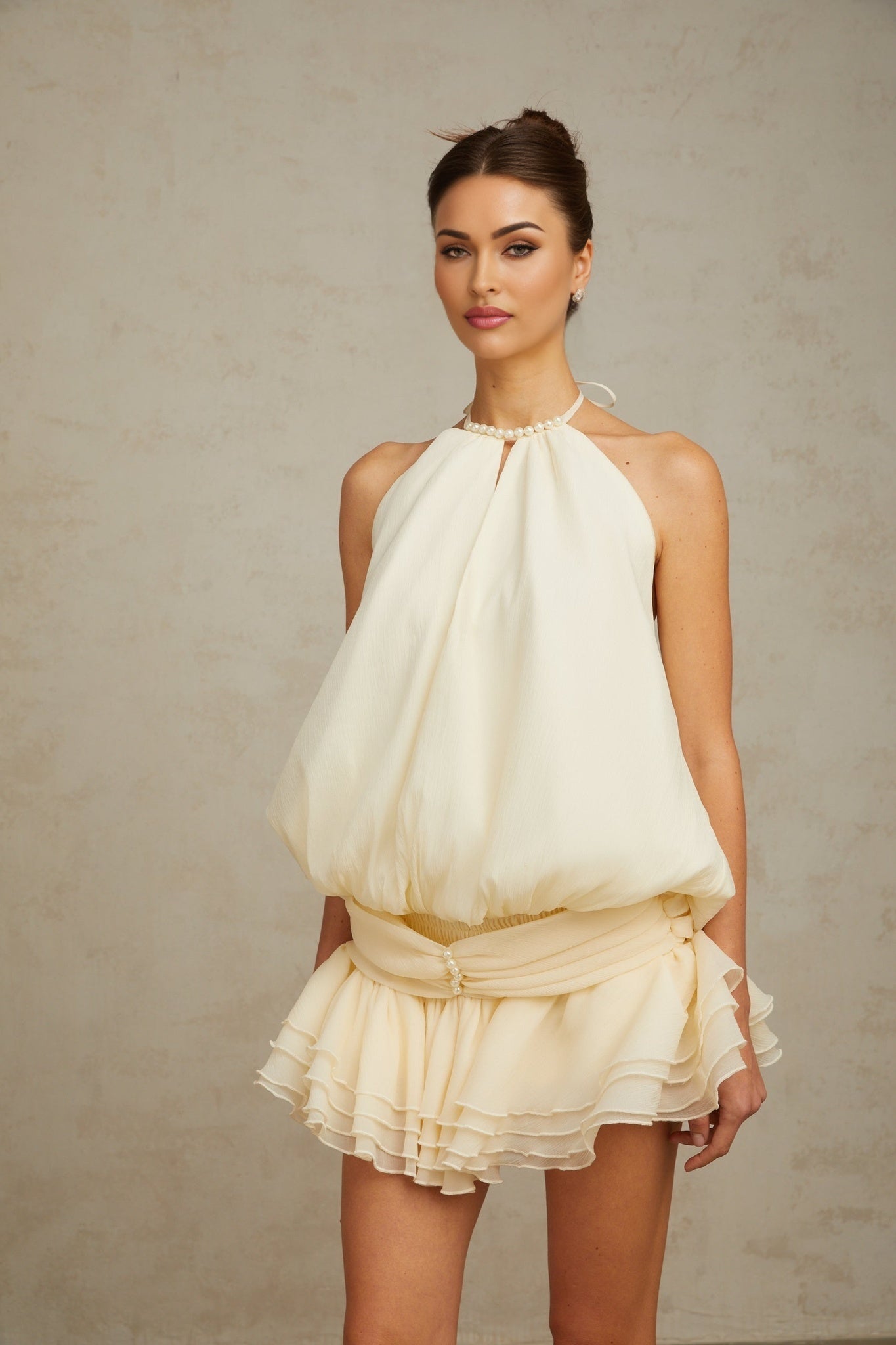 Young woman wearing a short white halterneck dress adorned with faux pearls standing in a neutral pose against a plain background