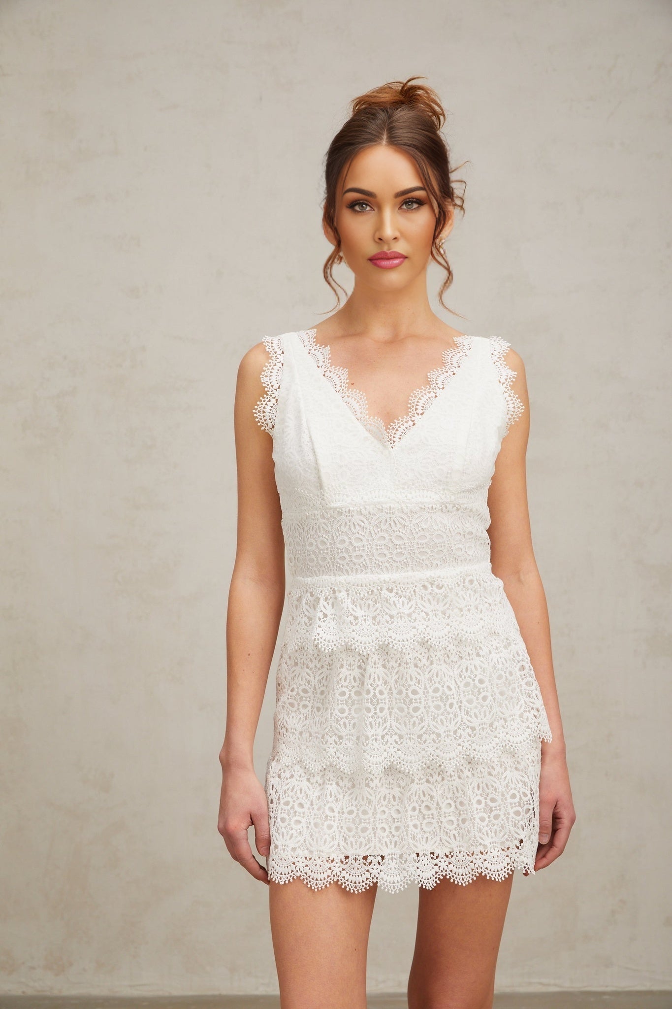 Young woman wearing a white lace mini dress with tiered ruffles and short sleeves standing against a plain background