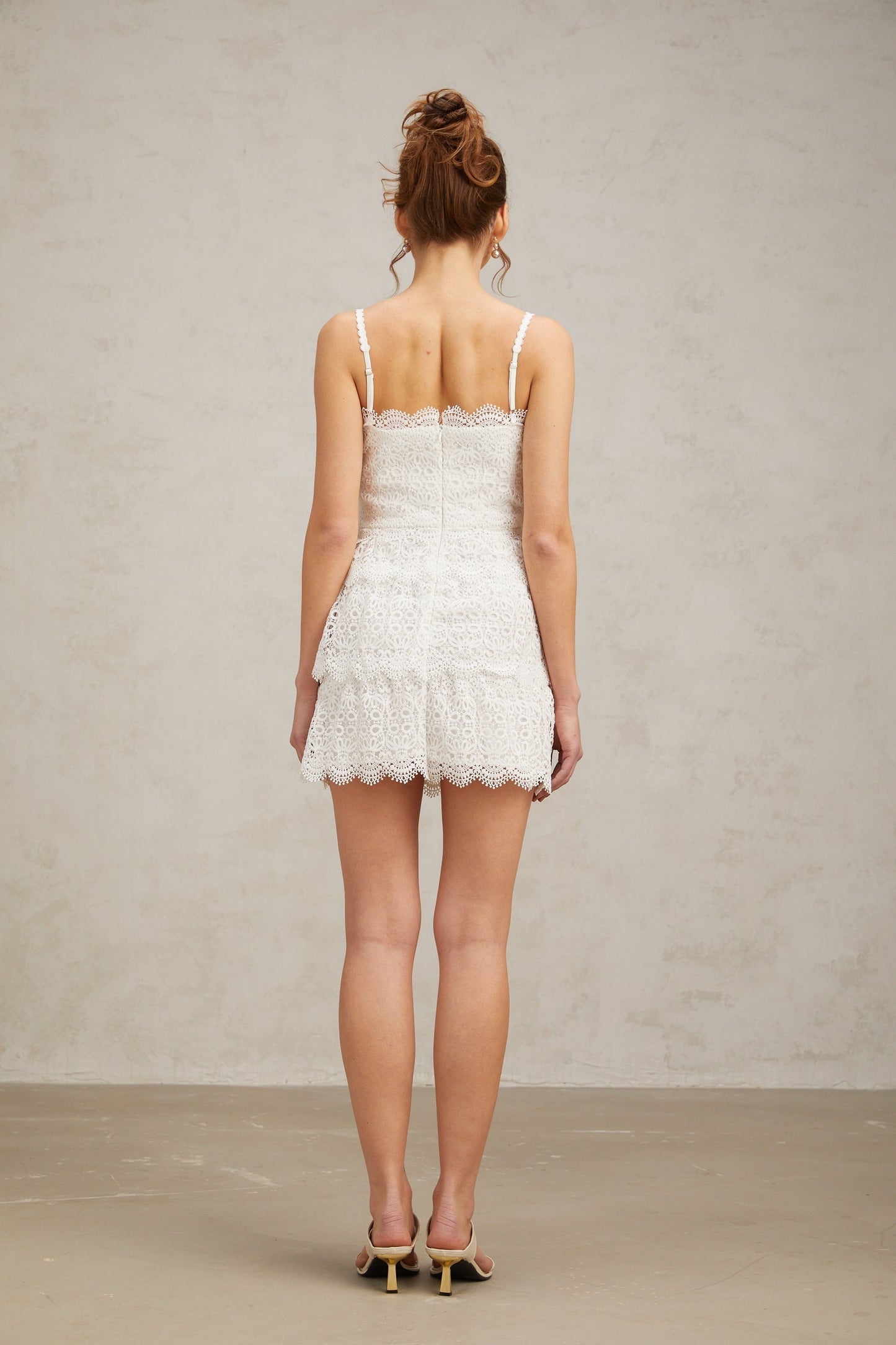 Young woman wearing a white lace mini dress with tiered skirt and short sleeves standing against a plain background