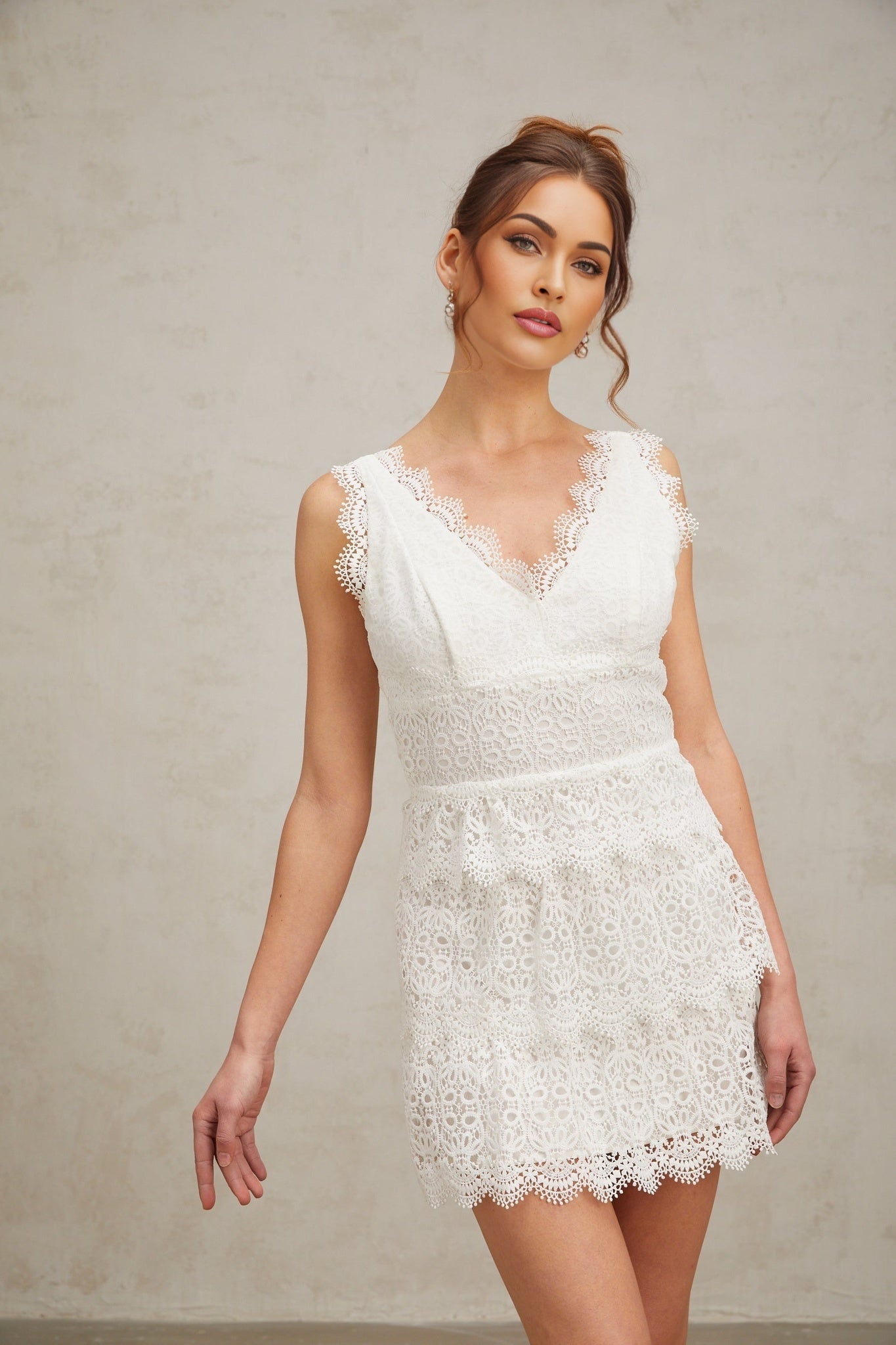 Young woman wearing a white lace mini dress with tiered skirt and short sleeves standing against a plain background