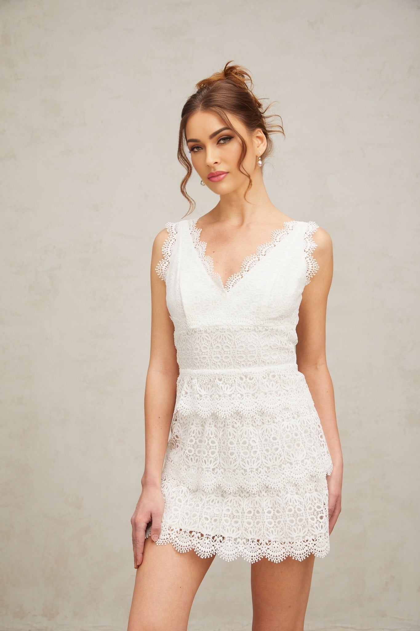 Young woman wearing a white lace mini dress with tiered skirt and short sleeves standing against neutral background
