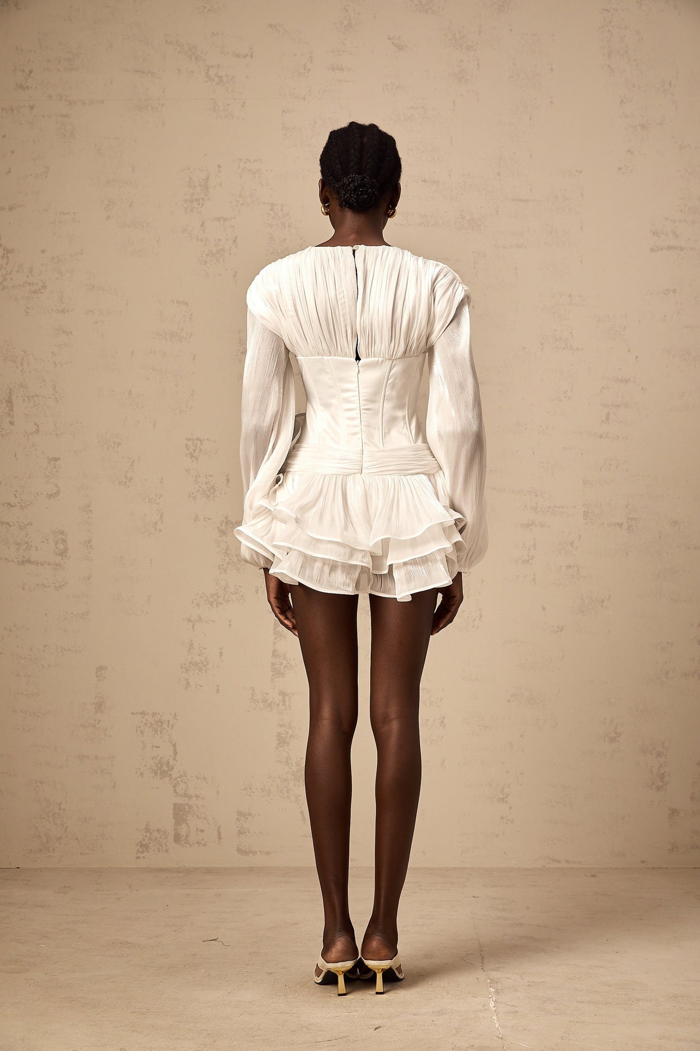 Young woman wearing a white mini dress with full pleats and cinched waist standing against plain background