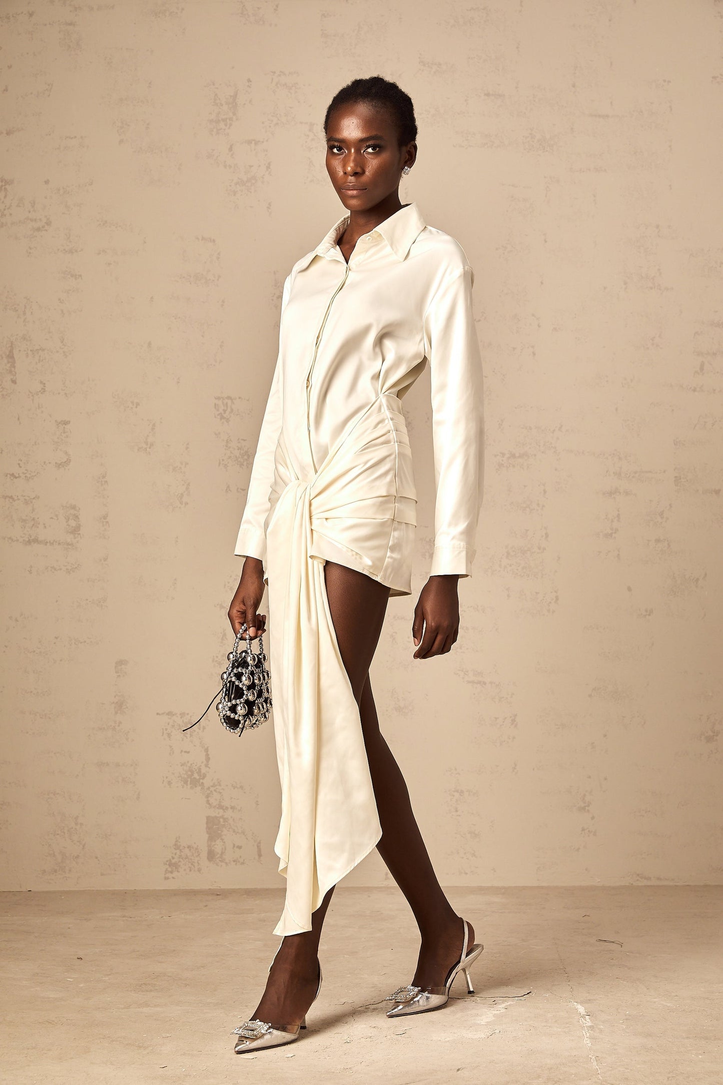 Young woman wearing a white mini shirt dress with ruched detailing and ribbon accents standing against a plain background