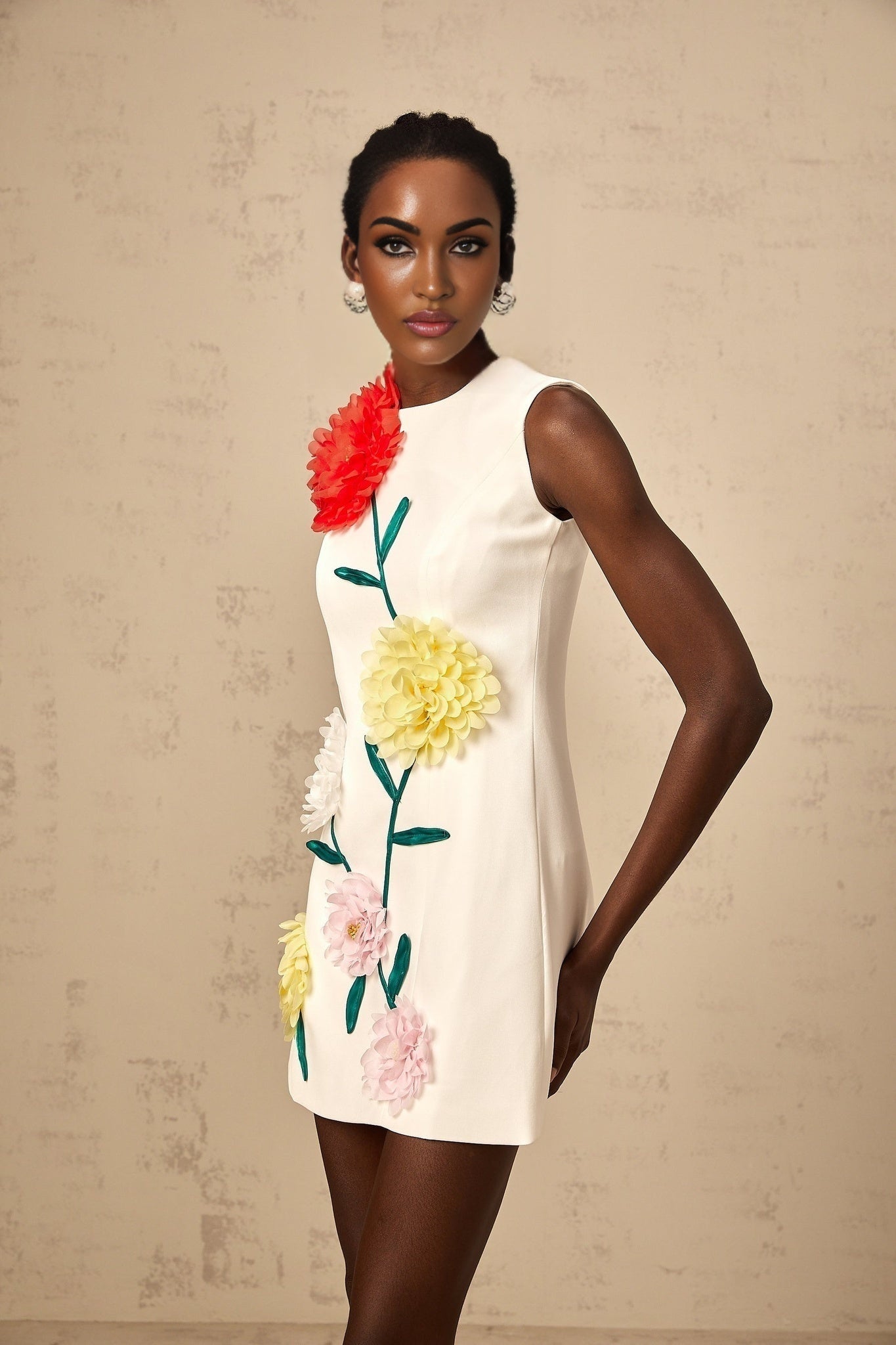 Young woman wearing a white sleeveless mini dress with three dimensional floral petal details standing against a plain background