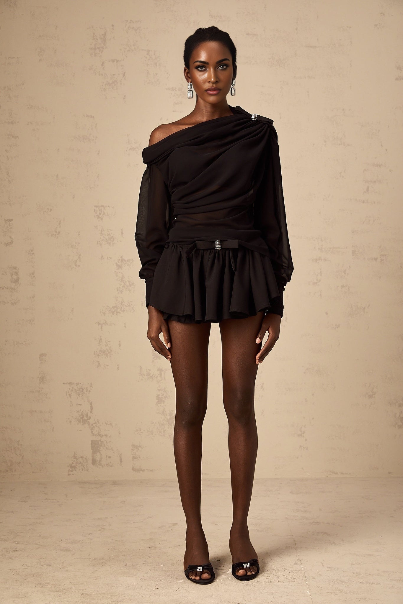 Young woman wearing black asymmetric tulle blouse and matching skirt set standing against plain background