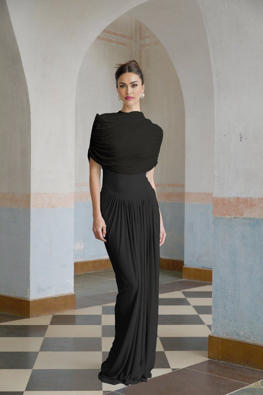 A black asymmetric pleated maxi dress worn by a model standing against a plain background The dress features a long flowing skirt with pleats and an asymmetric neckline or hemline