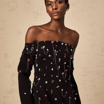 A black crystalembellished offshoulder maxi dress with a flowing silhouette worn by a model against a plain background