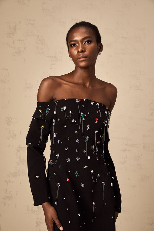 A black crystalembellished offshoulder maxi dress with a flowing silhouette worn by a model against a plain background