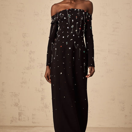 A black crystalembellished offshoulder maxi dress with a long flowing skirt worn by a model against a plain background