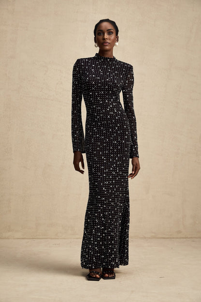 A black floor length maxi dress with sheer elements and decorative embellishments worn by a model