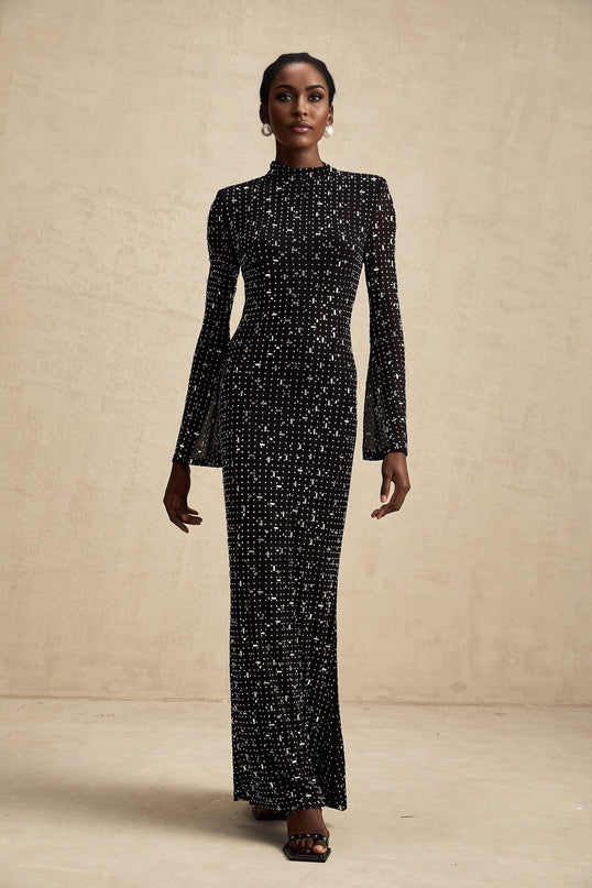 A black floor length maxi dress with sheer panels and embellished details worn by a woman standing in a neutral setting