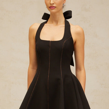 A black halterneck mini dress with a bow detail worn by a model standing against a plain background