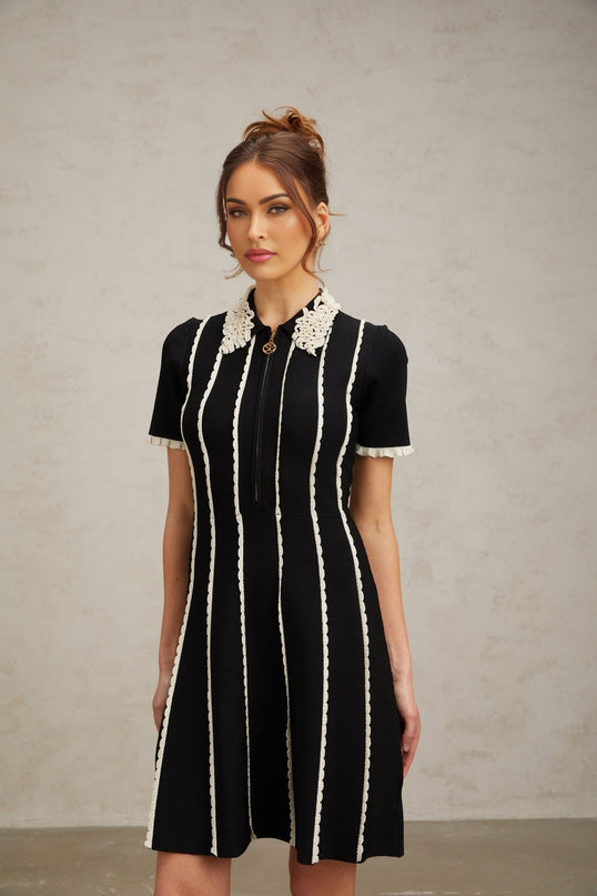A black knit mini dress with vertical white stripes and ruffle trim details worn by a model against a plain background