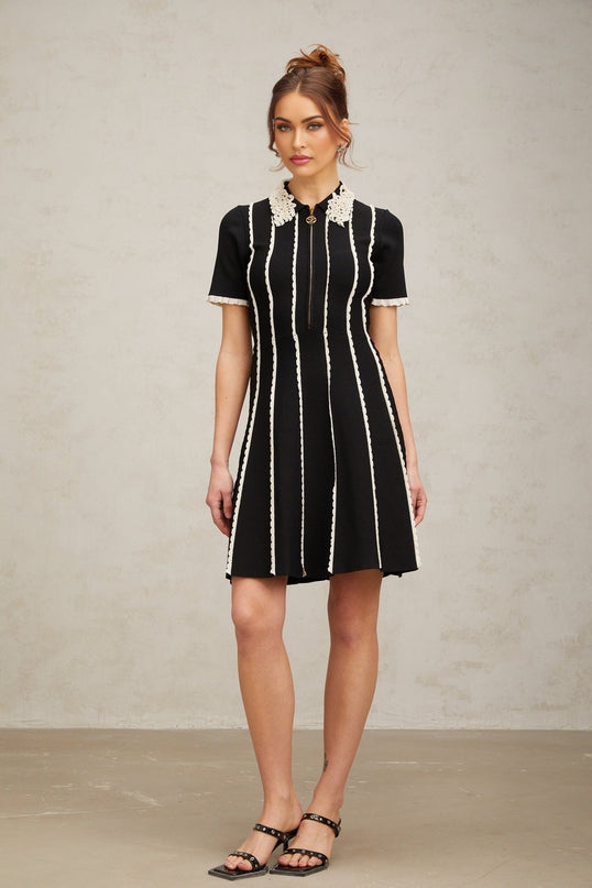 A black knit mini dress with vertical white stripes and ruffle trim details worn by a model against a plain background