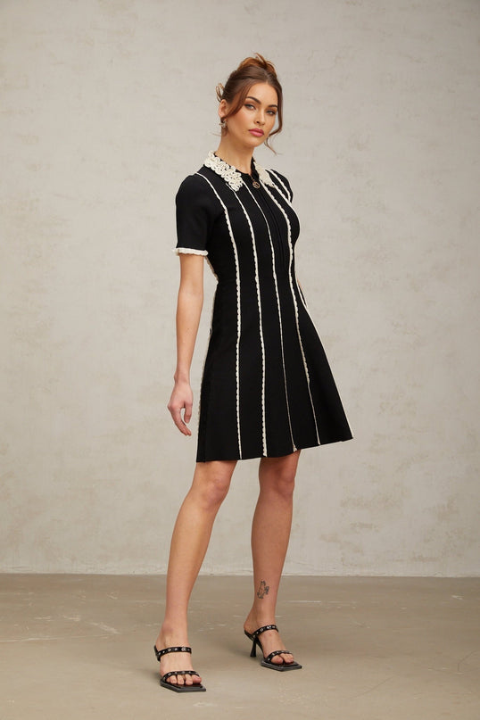 A black knit mini dress with vertical white stripes and ruffle trim details worn by a model standing against a plain background