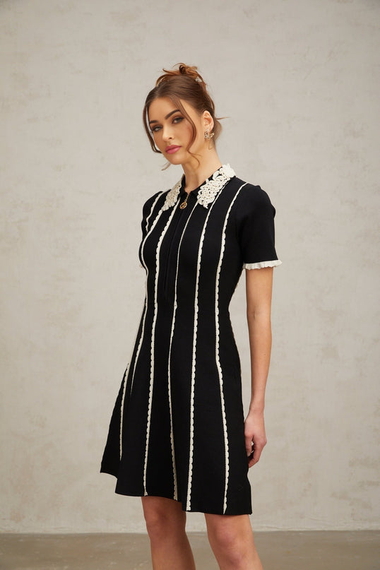 A black knit mini dress with vertical white stripes and ruffle trim details worn by a model standing against a plain background