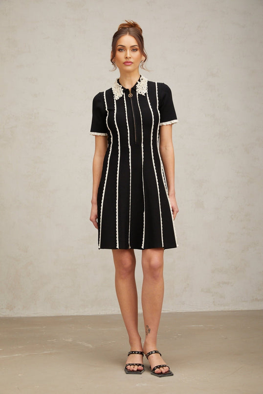 A black knit mini dress with vertical white stripes and ruffle trim details worn by a model standing against a plain background