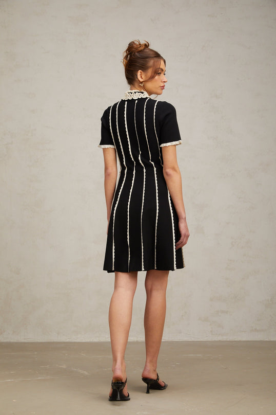 A black knit mini dress with vertical white stripes featuring ruffle trim details worn by a model against a plain background