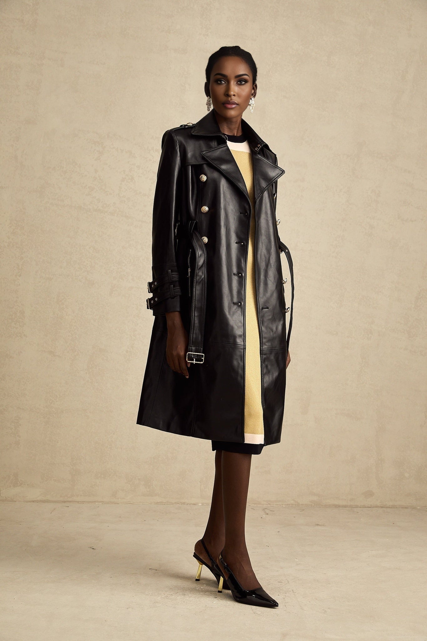 A black leather coat with double breasted front and belt at the waist worn by a model named Camille