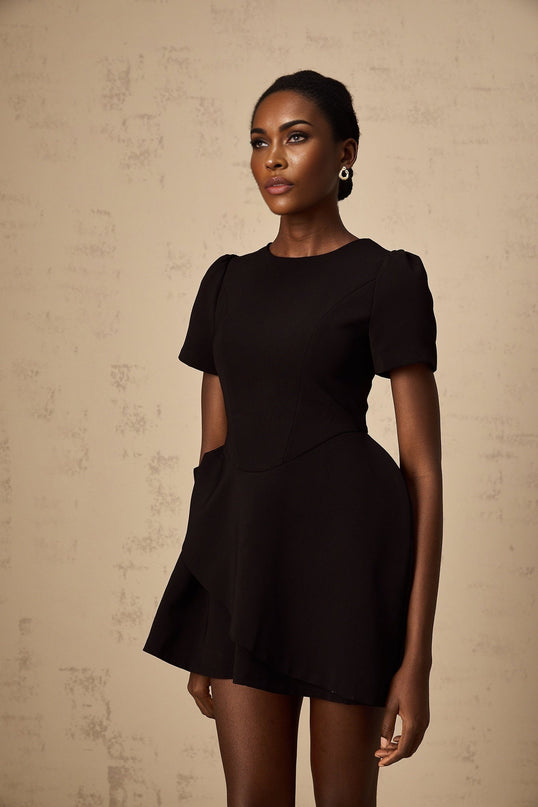 A black mini dress with asymmetric design and ruffled details modeled on a woman against a plain background