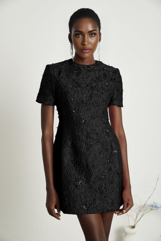 A black mini dress with floral applique embellishments featuring a short hemline and intricate flower details adorning the fabric