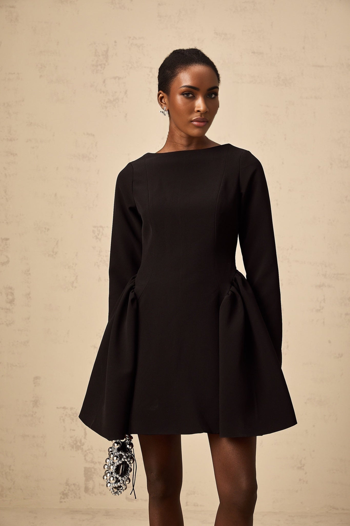 A black mini dress with long sleeves and a flared skirt worn by a model standing against a plain background