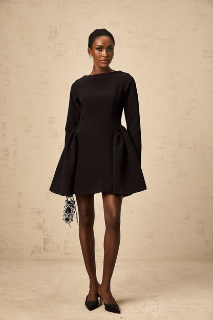 A black mini dress with long sleeves and a flared skirt worn by a model standing against a plain background