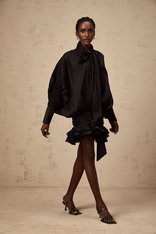 A black mini dress with ruffled details and a pussy bow neckline worn by a model named Melanie