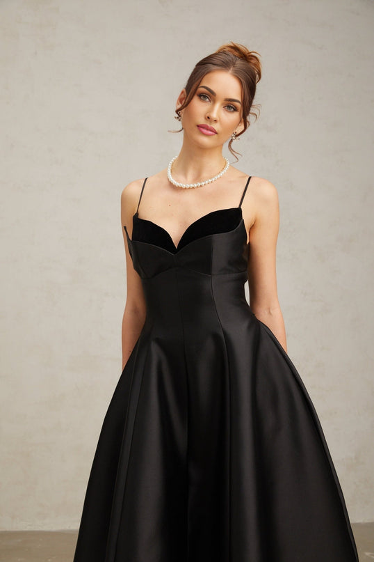 A black pleated midi dress with a flared skirt worn by a model standing against a plain background