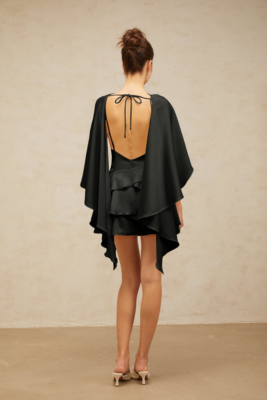A black satin mini dress with ruffled details and a shawl panel accent worn by a model against a plain background
