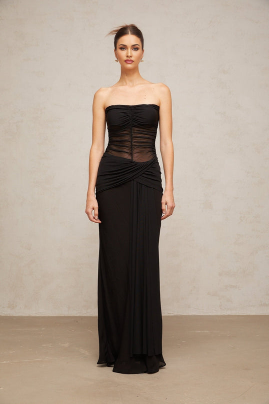 A black semi sheer maxi dress with pleated tulle fabric named Justine worn by a model standing in a neutral setting