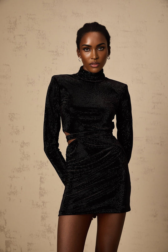 A black velvet mini dress with cut out details and stud embellishments worn by a model against a plain background