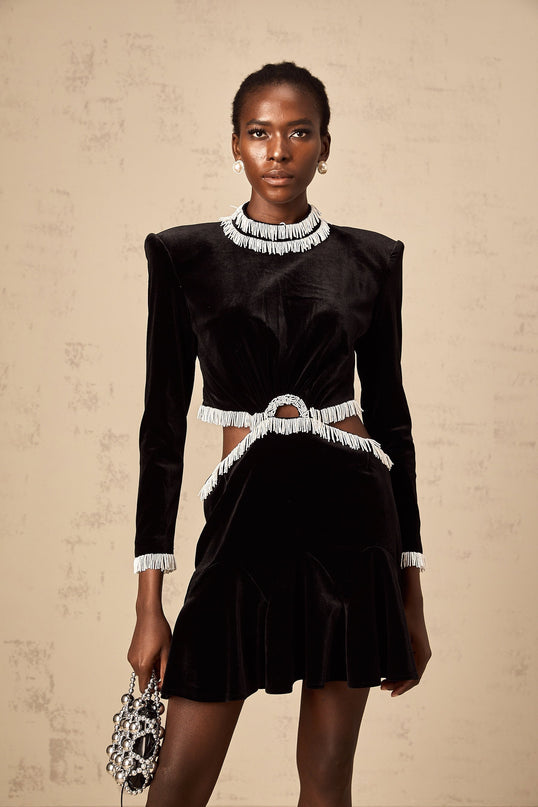 A black velvet mini dress with cutout details and fringe trim worn by a model standing against a plain background