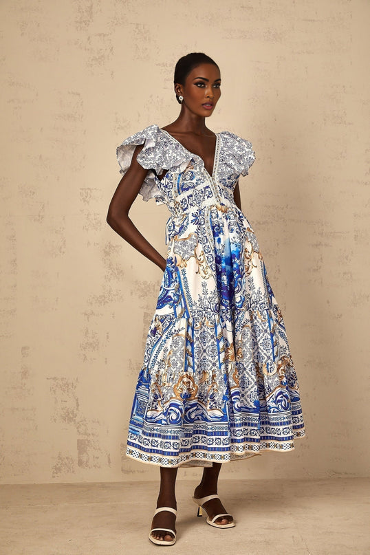 A blue midi dress with a baroque print pattern featuring tiered layers worn by a woman standing in a neutral background
