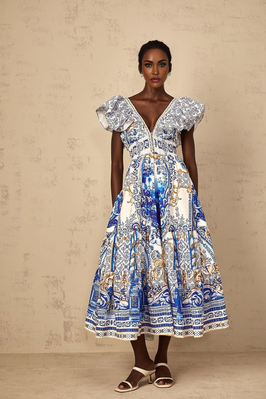 A blue midi dress with a baroque print pattern featuring tiered layers worn by a woman standing in a neutral background